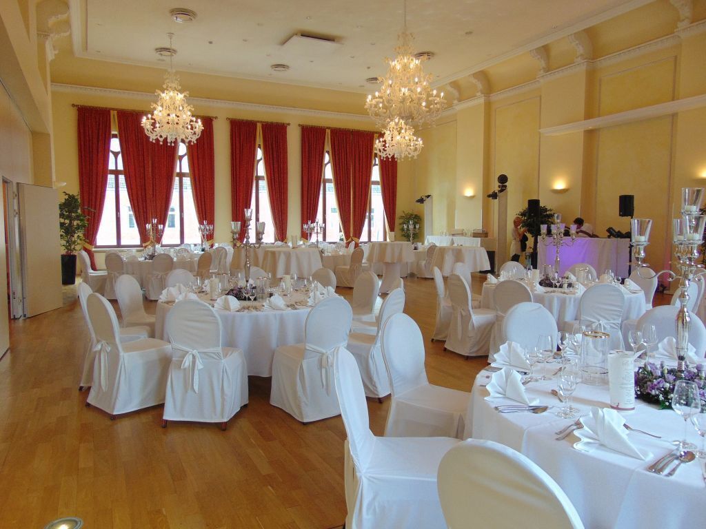 A banquet hall with round tables set for a formal event, featuring white tablecloths, chairs with white covers, and elegant chandeliers.