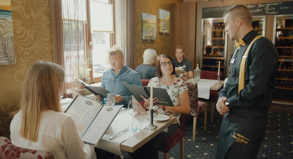 Interior de restaurante con comensales sentados en mesas, un camarero de pie y un menú sobre la mesa.