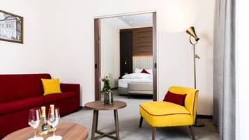 A hotel room with a red sofa, a yellow armchair, a small round table with two wine glasses, and a floor lamp.