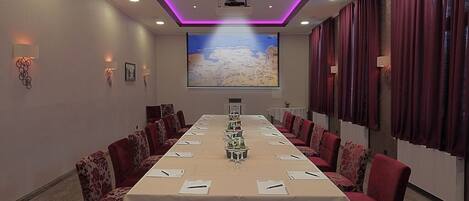 Une salle de conférence avec une longue table, des chaises à revêtement rouge, un grand écran et un éclairage d’ambiance violet.
