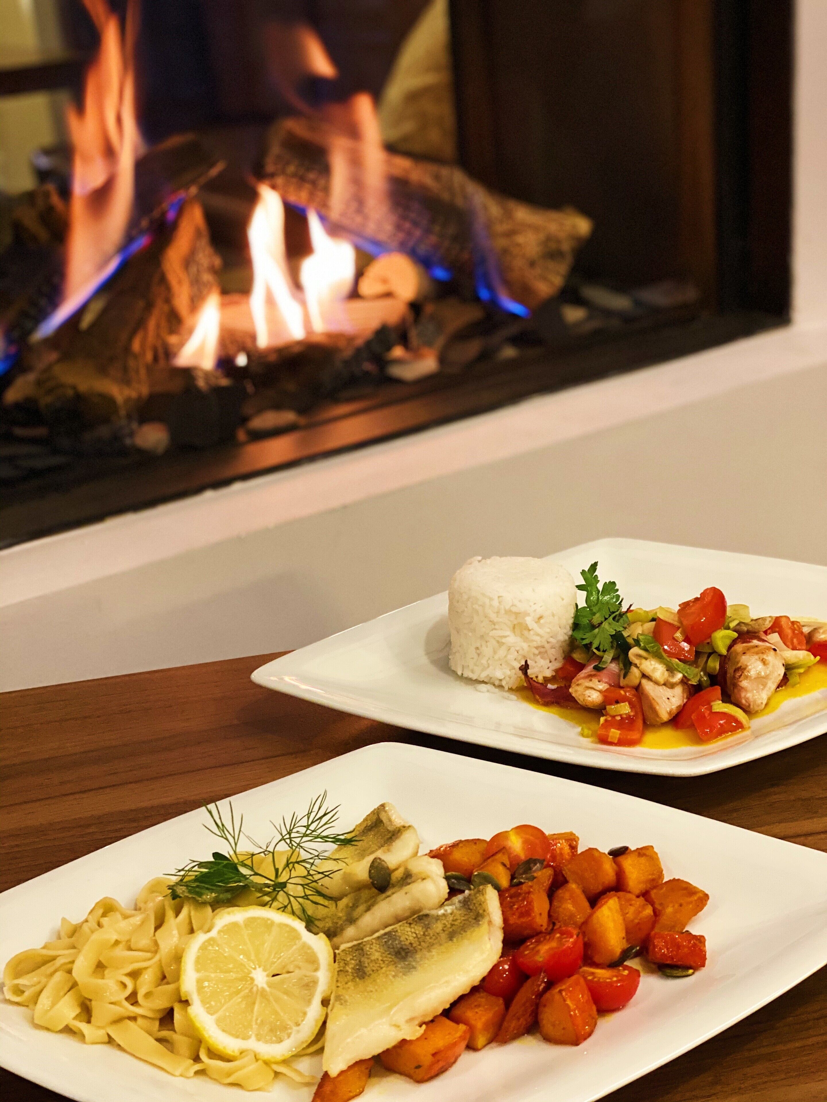 A dining table with two plates of food, one with rice and vegetables, and the other with pasta and fish, in front of a fireplace.