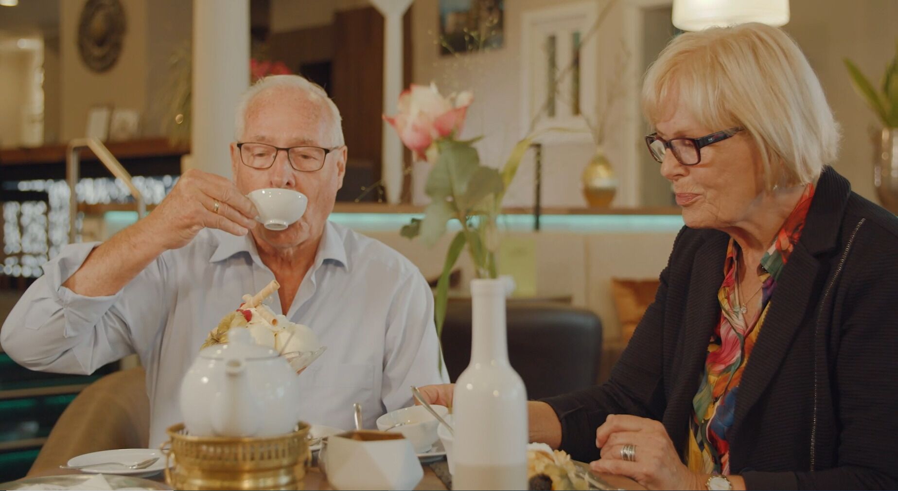 Två personer sitter vid ett bord med en tekanna, koppar och en vas med blommor.