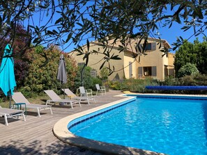 A swimming pool with lounge chairs and umbrellas, surrounded by greenery and a building.