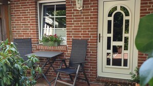 Een bakstenen huis met een pannendak, een witte deur en een raam. Buiten staat een tafel met twee stoelen.
