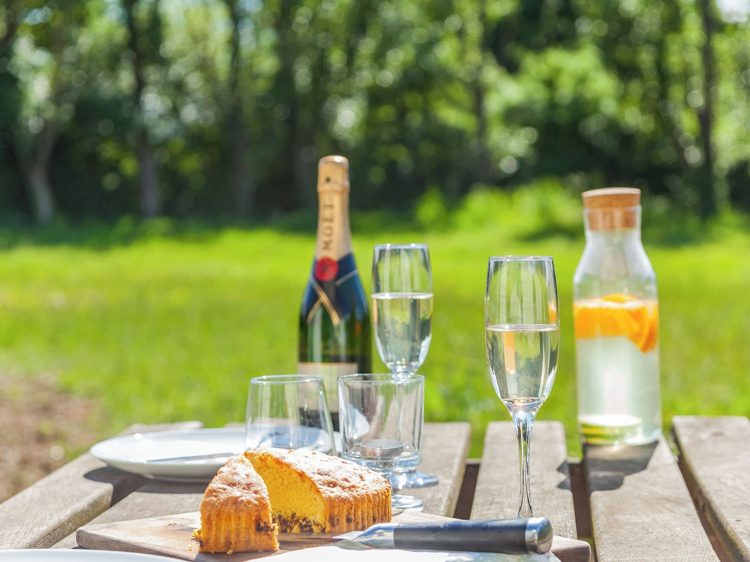 A picnic setup with a cake, champagne, and a bottle of lemonade on a wooden table.