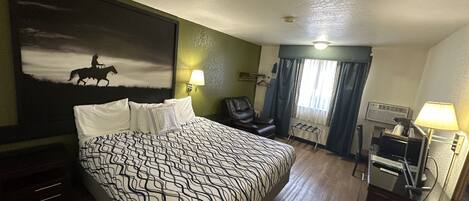 A hotel room with a large bed, a desk with a chair, a coat rack, a painting on the wall, and a window with curtains.