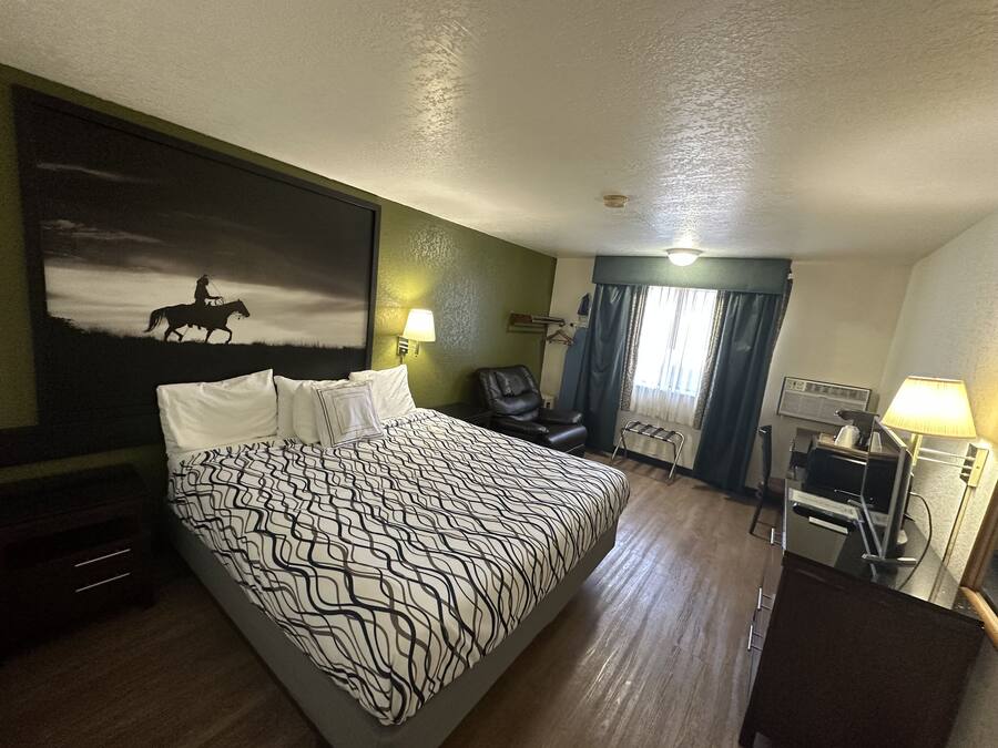 A hotel room with a large bed, a desk with a chair, a coat rack, a painting on the wall, and a window with curtains.