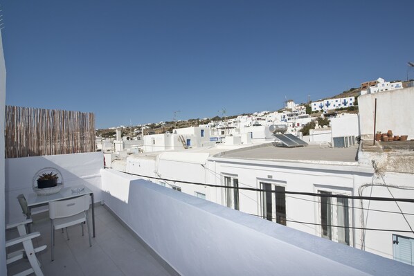 A rooftop terrace with a white table and chairs, a potted plant, and a view of white buildings in the distance. - Karboni Hotel (Mykonos)