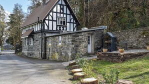 A traditional stone building with a black and white exterior, a small attached structure, and a gravel path with wooden stumps. - Crogen Gatehouse Lodge (Corwen)