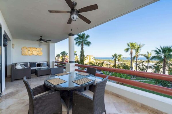 A covered outdoor seating area with a round table and chairs, a ceiling fan, and a view of palm trees and buildings.