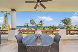 A covered outdoor seating area with a table and chairs, a ceiling fan, and a view of the ocean and palm trees.