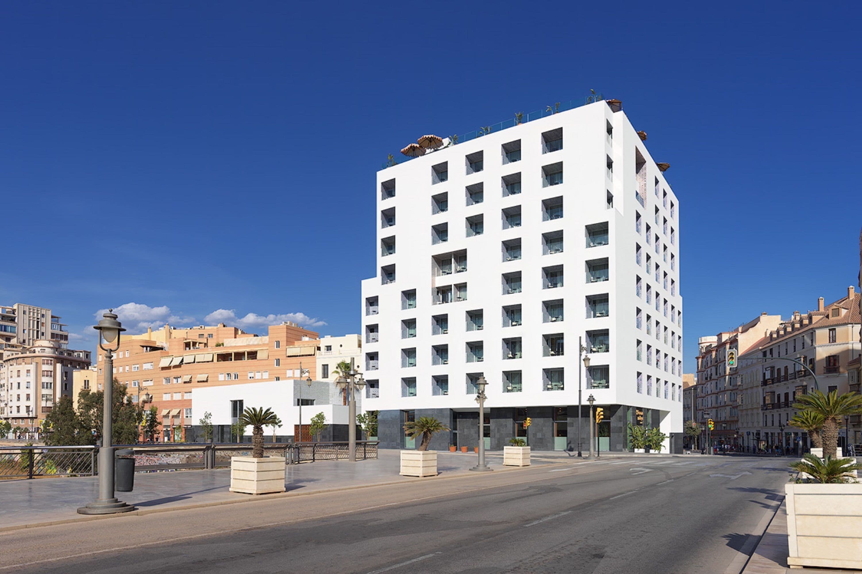 Un moderno edificio blanco de varios pisos y balcones, ubicado en una calle de la ciudad con palmeras y otros edificios al fondo.