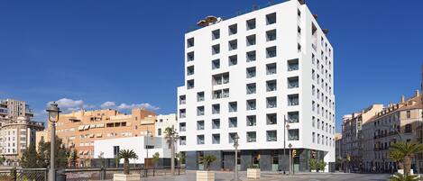 Un moderno edificio blanco de varios pisos y balcones, ubicado en una calle de la ciudad con palmeras y otros edificios al fondo.
