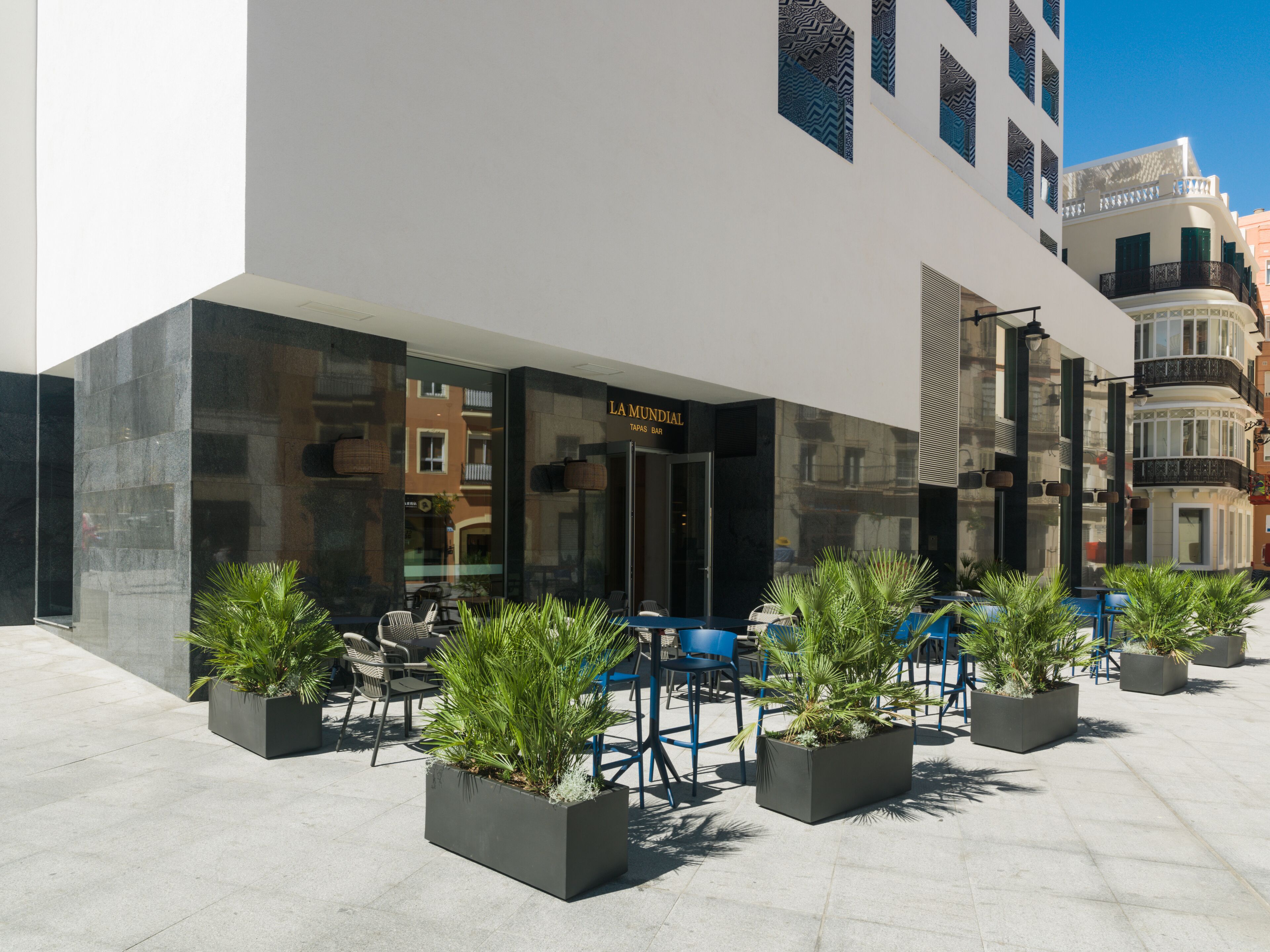 Een modern gebouw met een glazen gevel, een terras met stoelen en plantenbakken, en een bordje met de tekst 'LAMUNDIAL TAPAS BAR'.