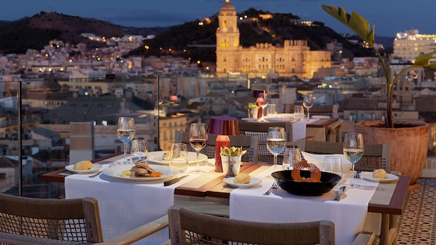 Un área de comedor en la azotea con mesas dispuestas para dos, con vistas a un paisaje urbano que incluye edificios históricos y una colina al fondo.