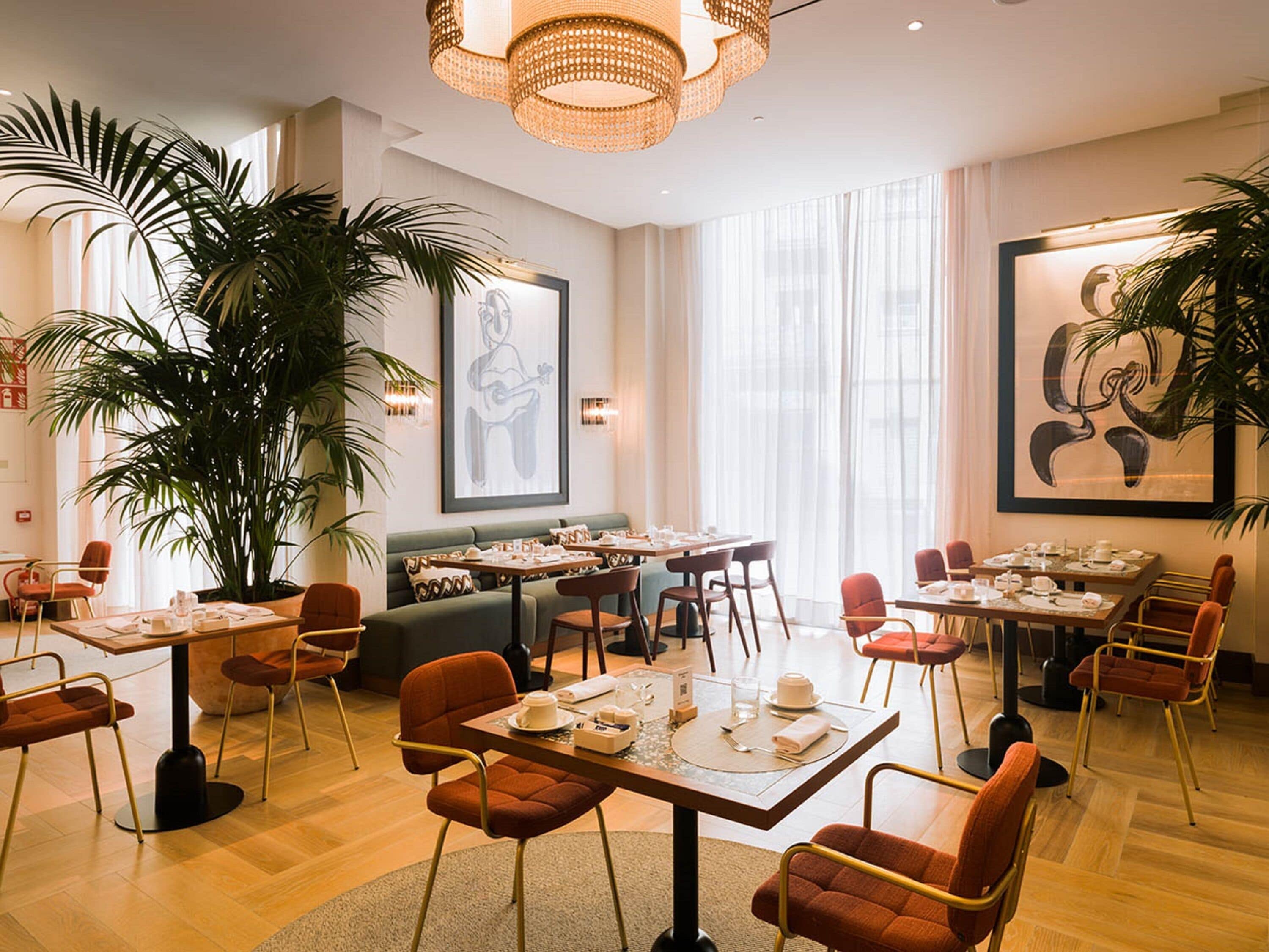 A dining area with tables set for service, large potted plants, and artwork on the walls.