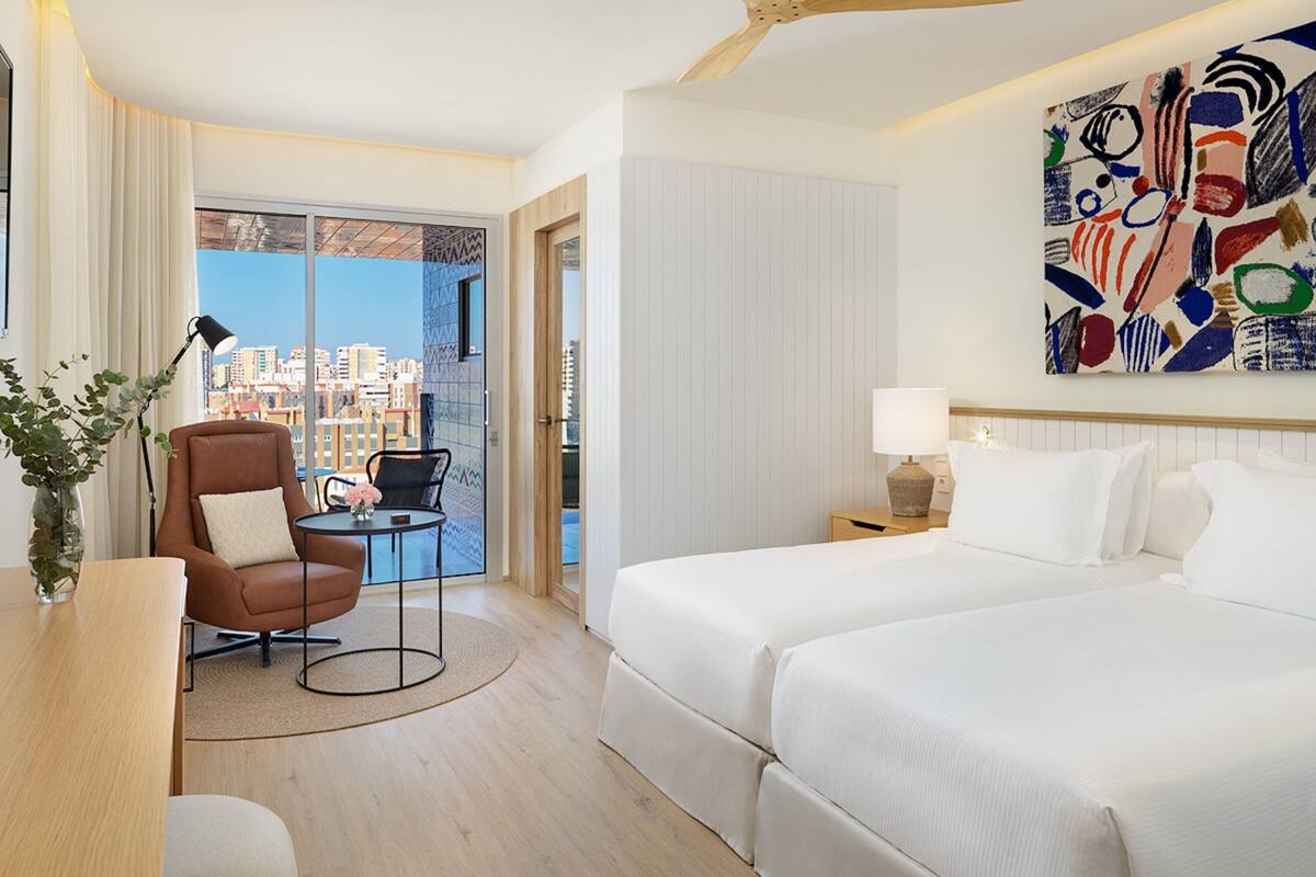 Habitación de hotel con cama, escritorio, silla, lámpara y vista a la ciudad a través de un ventanal grande.