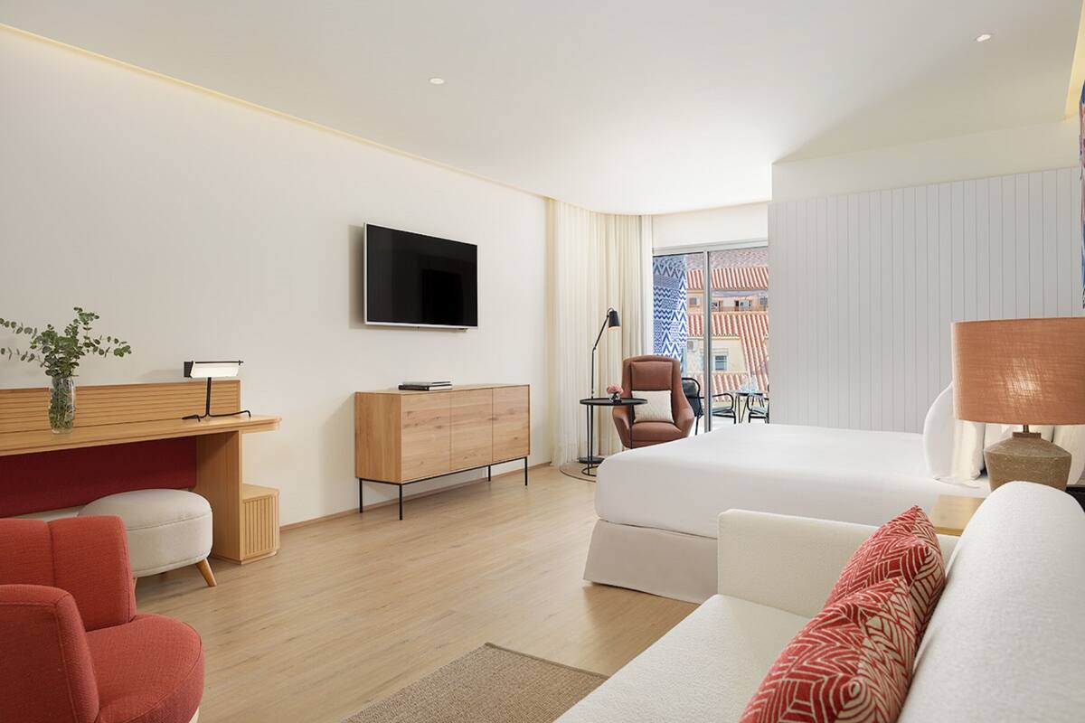 Una habitación de hotel moderna con una cama, un escritorio con televisor, una silla roja y vistas a edificios a través de una ventana.