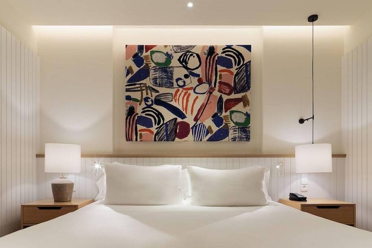 Habitación de hotel con cama, dos mesitas de noche, una lámpara, un cuadro en la pared y una lámpara colgante.