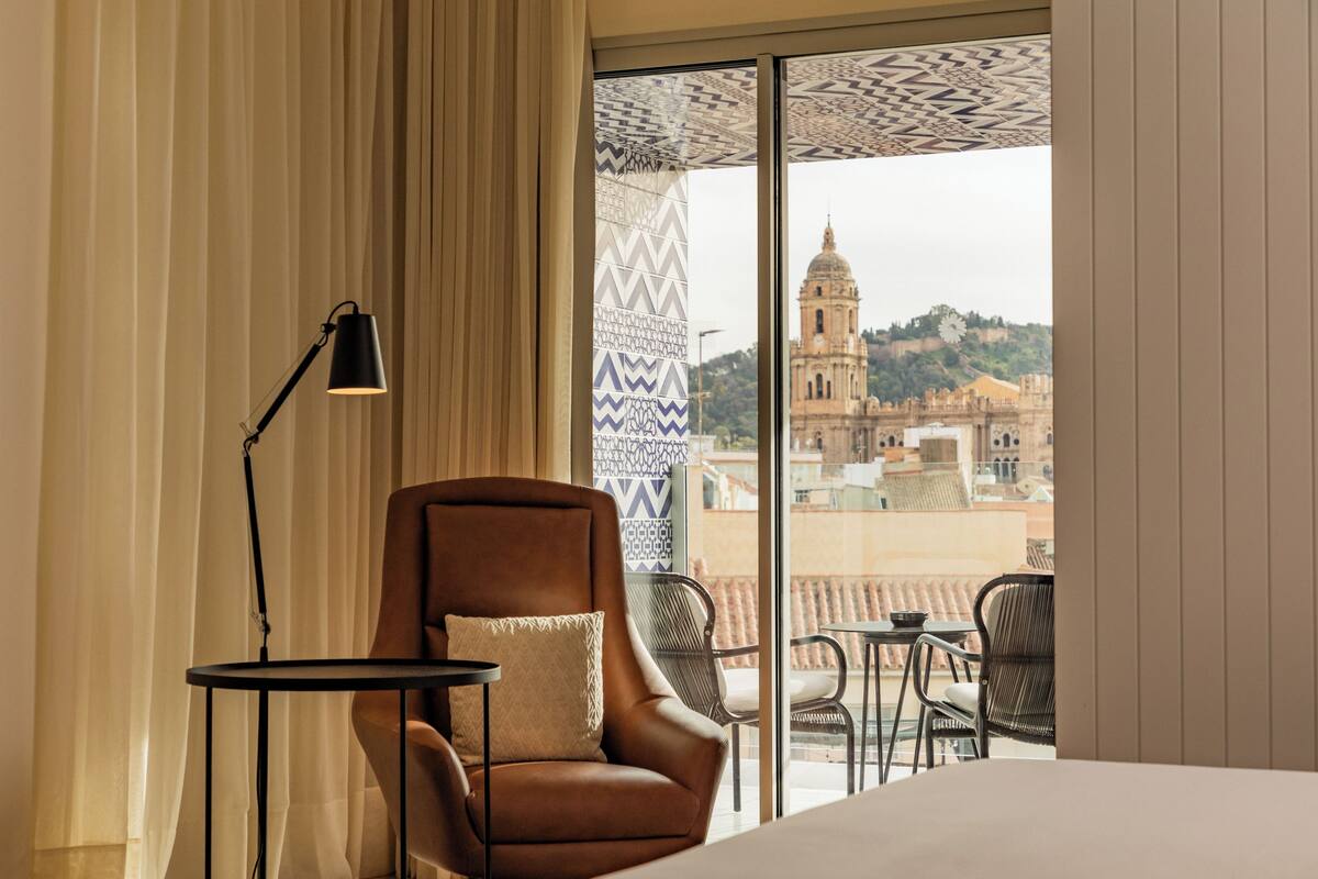 Habitación de hotel con un ventanal grande, un sillón de cuero marrón, una mesita pequeña y una lámpara.