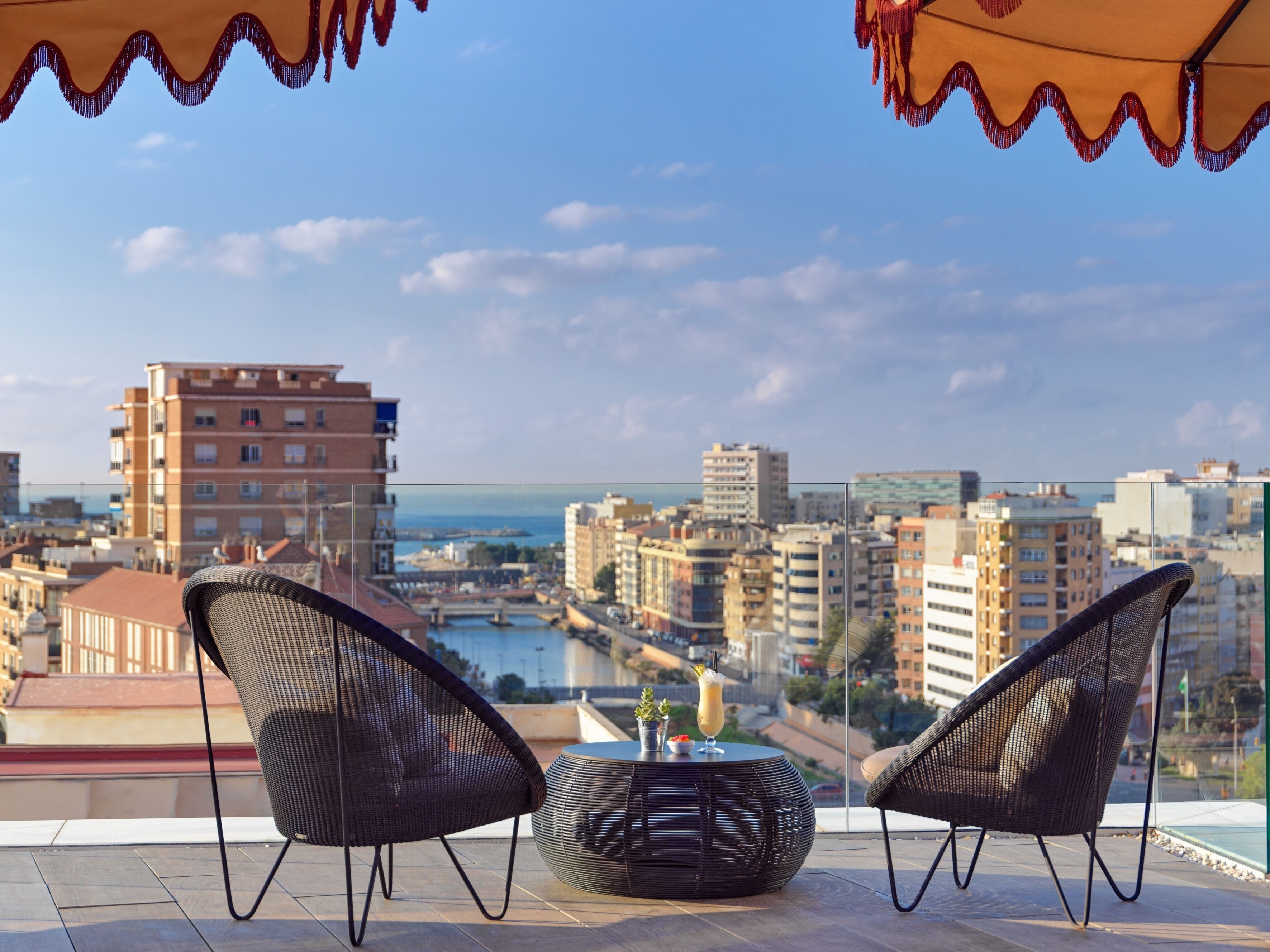 A balcony with wicker chairs and a round table, overlooking a cityscape with buildings and a body of water.