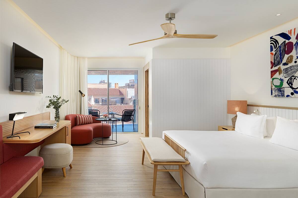Una habitación de hotel con una cama, un escritorio, un sofá rojo, una mesita, una silla, una lámpara de pie y un ventilador de techo.