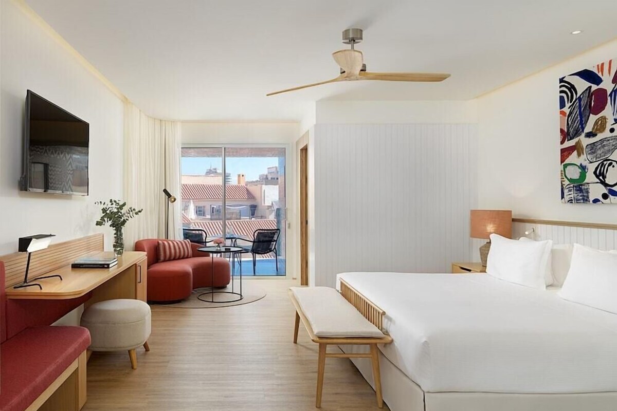 Habitación de hotel con cama, escritorio, sofá, silla, mesita, planta, ventilador de techo, ventana y un cuadro en la pared.