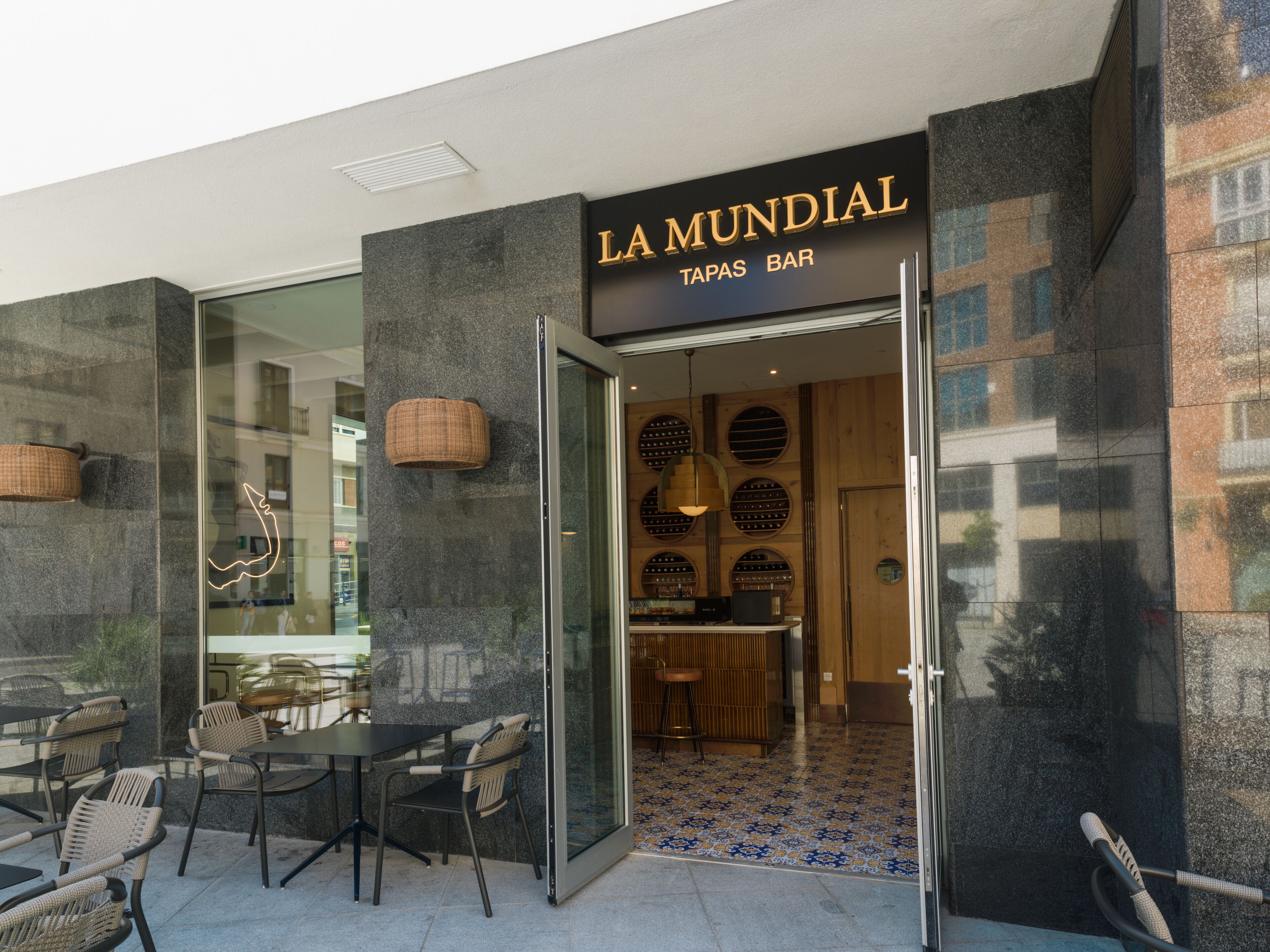 El exterior del bar de tapas La Mundial, con un diseño moderno, que cuenta con una entrada de cristal, zona de estar al aire libre y un letrero sobre la puerta.