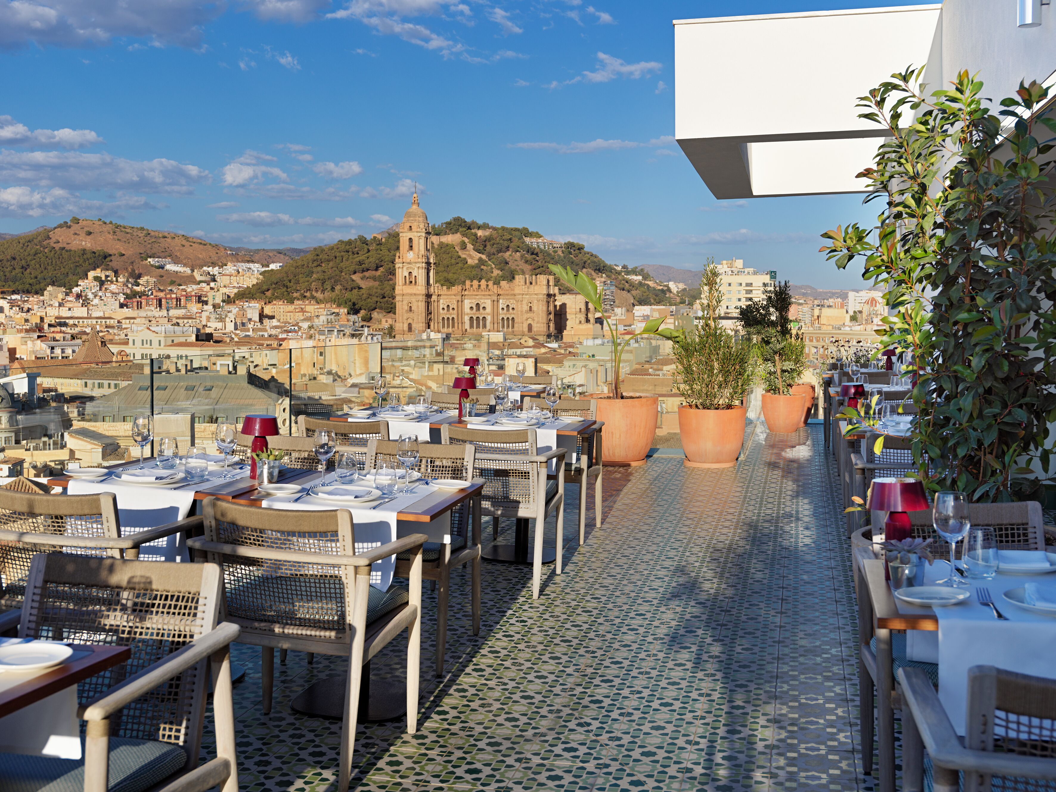 Una terraza en la azotea con mesas dispuestas para comer, plantas en macetas y vista a un edificio histórico al fondo.