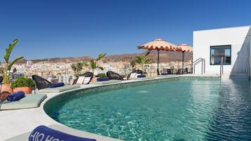 Zona junto a la piscina con sillones, sombrillas y vista a un paisaje urbano al fondo.