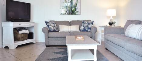 A living room with a grey sofa set, a white coffee table, a flat-screen TV on a white stand, a framed plant on the wall, and a lamp on a side table.