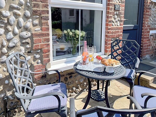 An outdoor patio with a round table, two chairs, and a window with a view of a garden. - 2 bedroom accommodation in Sheringham (Sheringham)