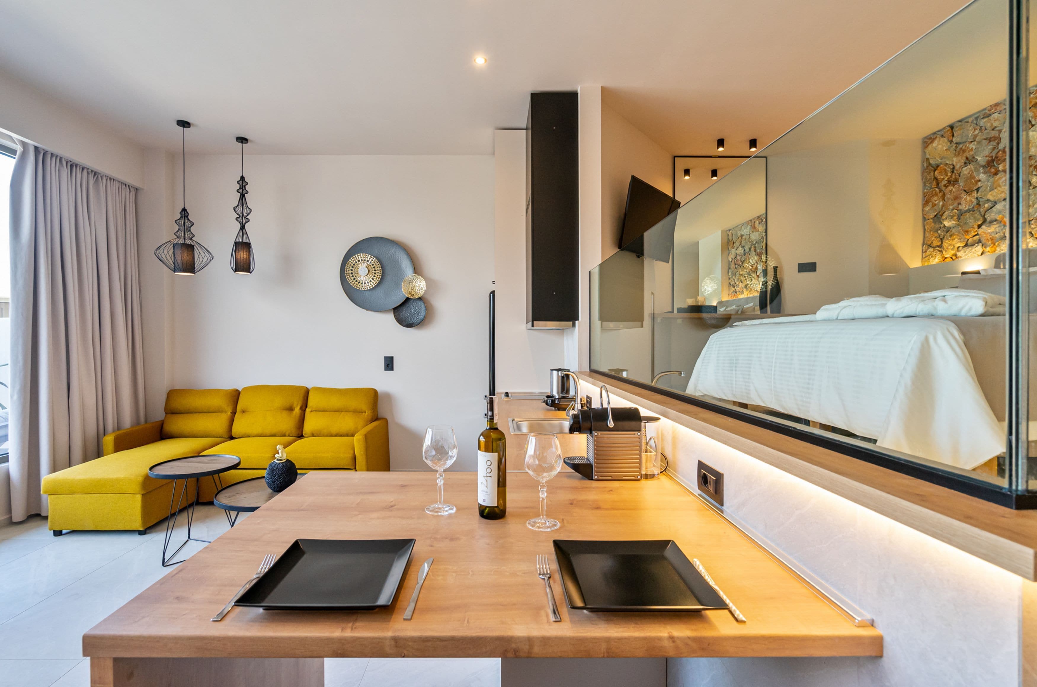 Habitación de hotel moderna con una mesa de comedor de madera dispuesta para dos personas, un sofá amarillo, un espejo grande y vista a la cama.