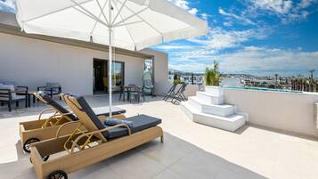 Terraço no telhado com cadeiras de descanso, um grande guarda-sol e vista para uma piscina e edifícios ao longe.