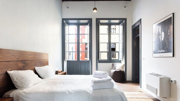 Un dormitorio con cabecera de madera, una cama grande con ropa de cama blanca, dos ventanas con marcos negros, un radiador y un cuadro enmarcado en la pared.