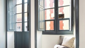 Una habitación con suelo de madera, un ventanal con vistas a edificios y un sillón informal de color marrón.