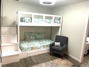 A bunk bed with a patterned bedspread and pillows, a gray armchair with a 'welcome to our beach house' pillow, and a patterned rug on a wooden floor.