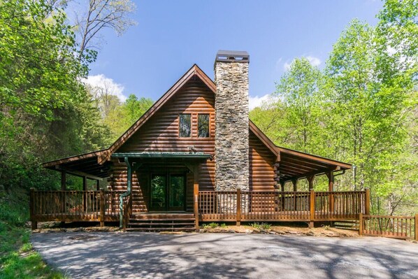 A log cabin with a stone chimney, wooden porch, and a fenced driveway.