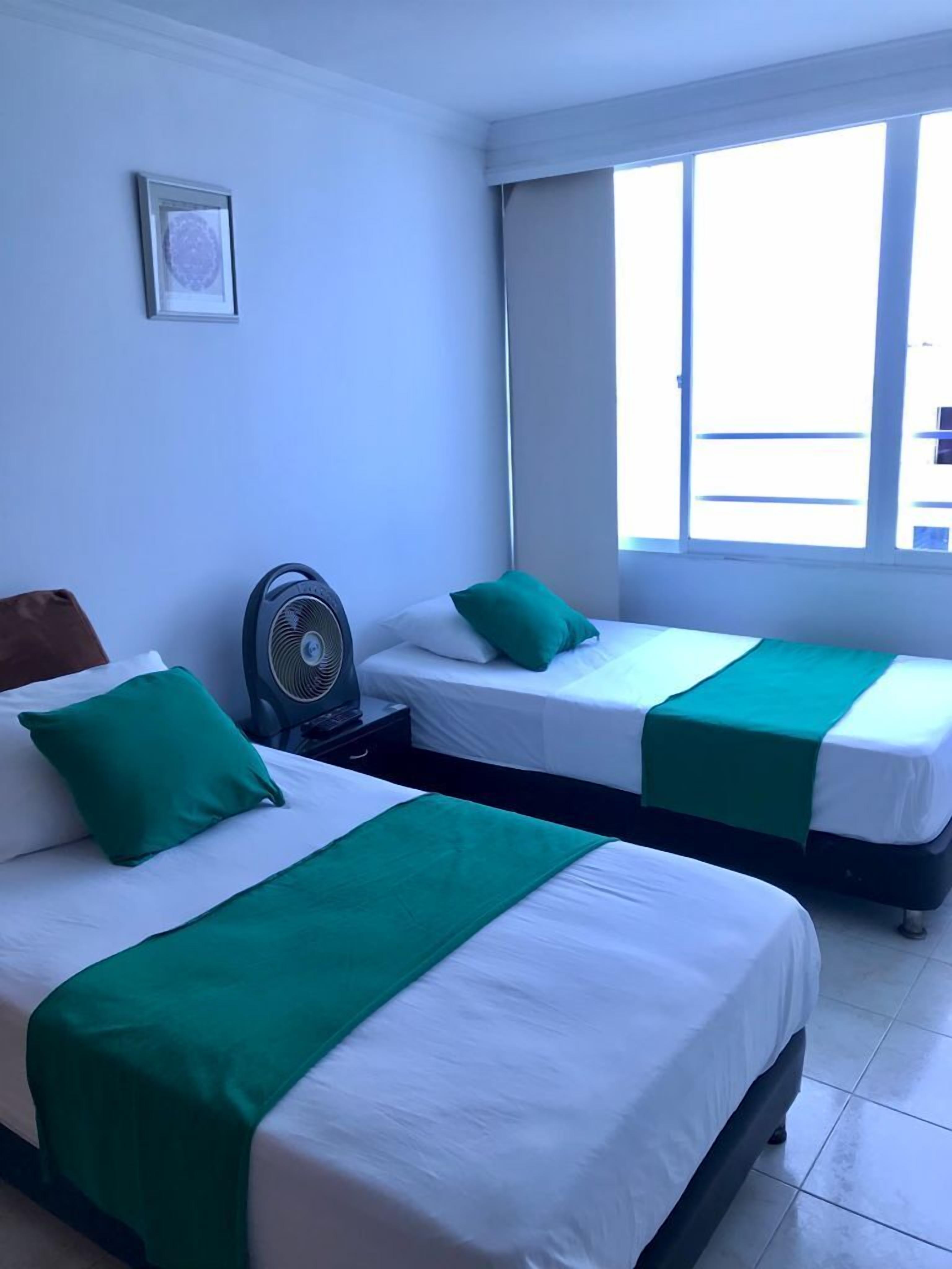 A hotel room with two beds, each with green and white bedding, a standing fan, and a window providing a view of the outside.
