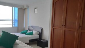 A hotel room with a large wardrobe, two beds with green bedding, a small bedside table, and a window with a view. - Apto VISTA AL MAR piso 16 PLAYA CERCA (Cartagena)