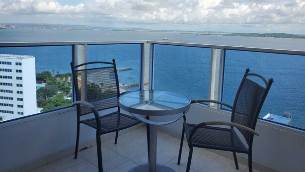 A balcony with a glass table and two chairs, overlooking a body of water and distant land. - Apto VISTA AL MAR piso 16 PLAYA CERCA (Cartagena)