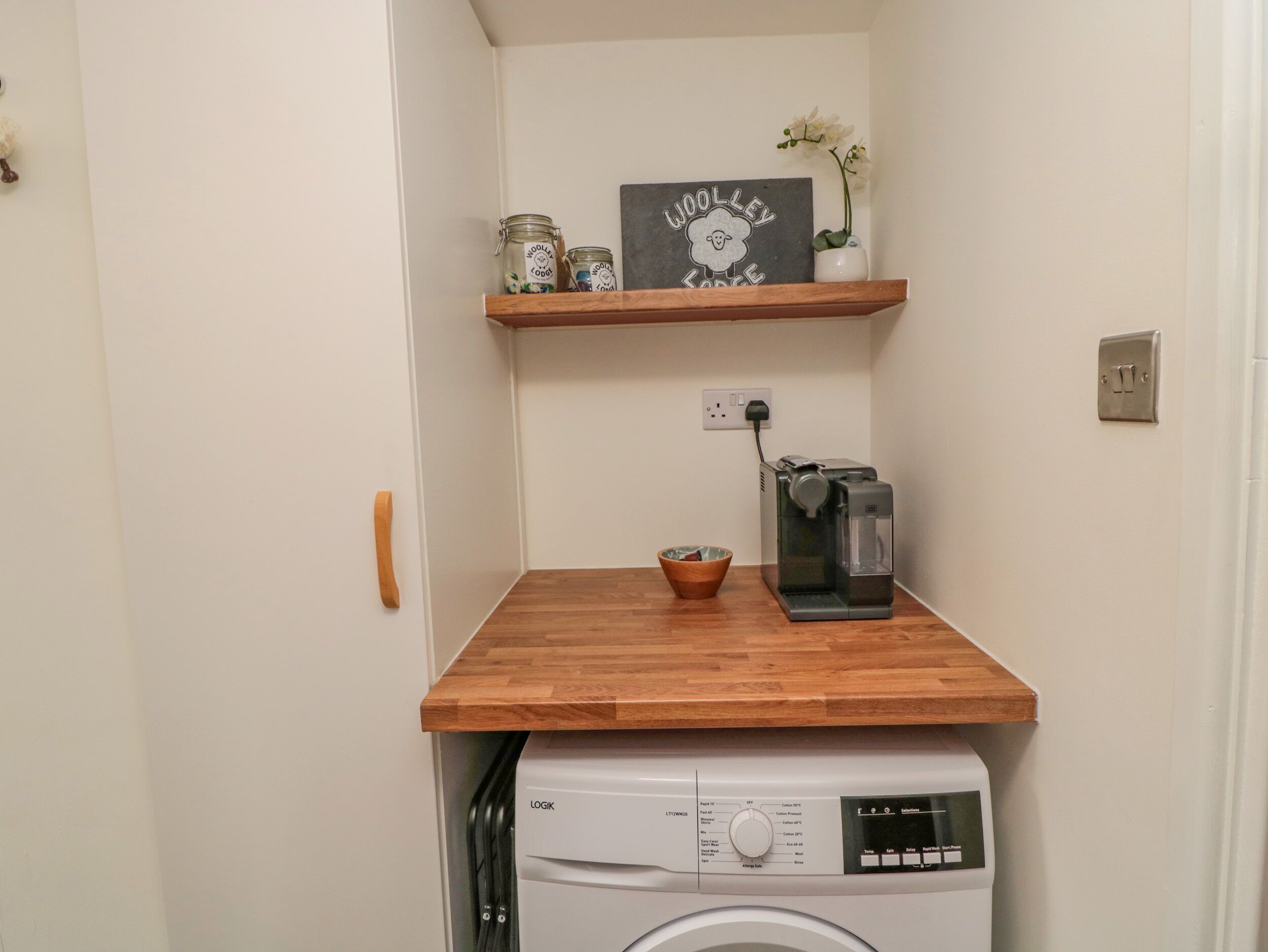 Een ruimtelijke wasruimte met een wasmachine, een koffiezetapparaat en een plank met potten en een decoratief bord.