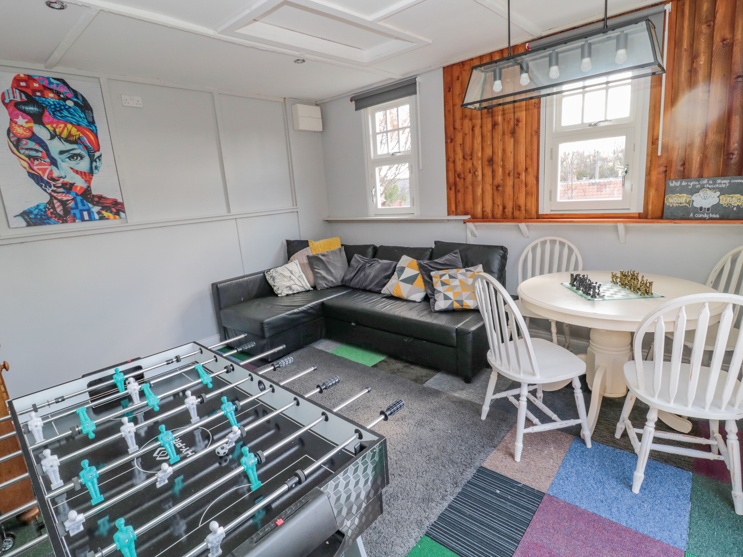 Una habitación con mesa de futbolín, sofá, mesa de comedor con sillas y ventana con paneles de madera.