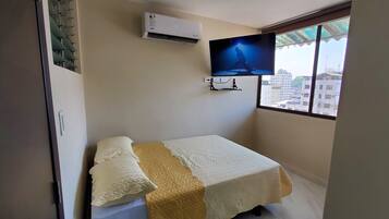 A room with a bed, a mounted TV, an air conditioner, and a window with a view of buildings.