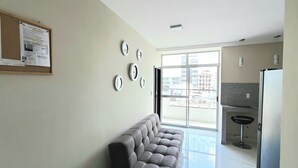 Un salon moderne comprenant un canapé, un tableau d’affichage avec des avis, une grande fenêtre et un coin cuisine avec un réfrigérateur et un tabouret de bar.
