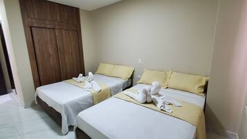Habitación de hotel con dos camas, cada una con cubrecama blanco y manta color beige. Se observa un armario de madera y una puerta.