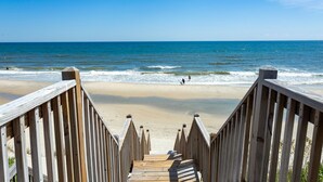 Ein Holzsteg führt zu einem Strand mit Wellen und blauem Himmel.