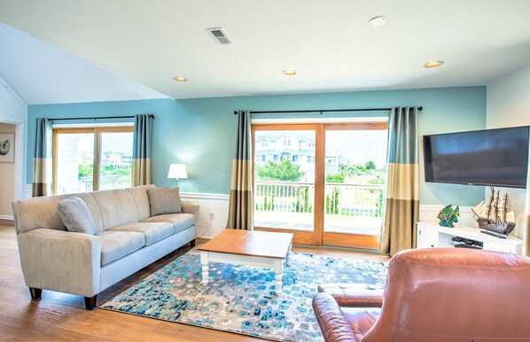 A living room with a sofa, coffee table, and ottoman, a flat-screen TV, and a balcony with a view of houses.