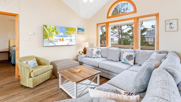 A living room with a large gray sectional sofa, a small green armchair, a wooden coffee table, and a ceiling fan. There are two windows with wooden frames and a view of the outside. - It's 5 O'Clock Somewhere – Beach,  Pool & Hot Tub (Corolla)