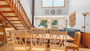 Un comedor con sillas de madera y una mesa, una escalera, un sofá y un ventanal con cortinas.